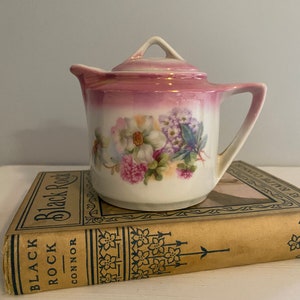 May include: A small, pink and white porcelain creamer with a floral design. The creamer has a lid and a spout. It is sitting on top of a book titled "Black Rock" by Connor.