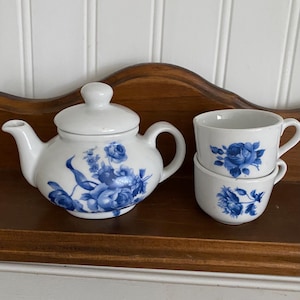 May include: White porcelain tea set with blue floral designs. The set includes a teapot with a lid and two teacups stacked on top of each other. The tea set is displayed on a wooden shelf.