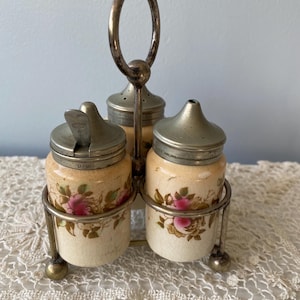 May include: A vintage salt and pepper set with three ceramic jars featuring pink rose designs, held in a silver-toned metal stand with a handle. The jars have metal lids and sit on a lace tablecloth.