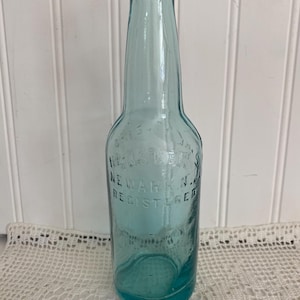 May include: A vintage, light blue glass bottle with embossed text that reads "Hensler's Newark N.J. Registered." The bottle has a long neck and a rounded base. The bottle is displayed on a white lace doily.