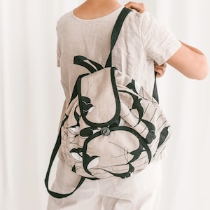 May include: A beige canvas backpack with black straps and a black floral print. The backpack has a flap closure with a button.