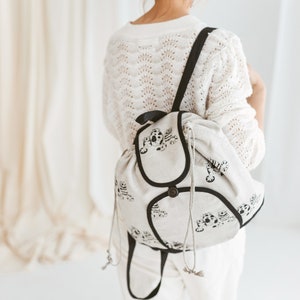 May include: A white canvas backpack with black trim and a drawstring closure. The backpack features a black and white print of stylized tigers.