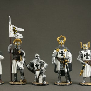 May include: A group of miniature medieval knight figurines. The figures are in detailed armour, with white and black crosses on their tunics. One figure holds a banner, and another a spear. The colour palette includes silver, white, black, and gold.