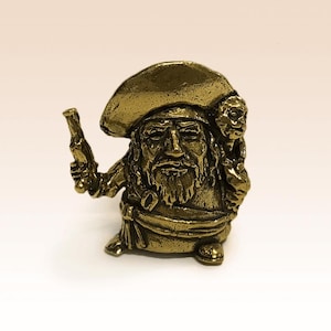 May include: A detailed brass figurine of a pirate, holding a pistol in one hand. The pirate wears a hat and has a long beard. A second face is sculpted on the back of the pirate's head. The figurine is a decorative item.