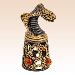 Op de afbeelding: Een decoratieve vingerhoed met een gebeeldhouwde kobrakop met een donkere metalen afwerking. De basis is een goudkleurig metalen rooster met een slangenschubstructuur, geaccentueerd door drie ronde, gepolijste barnsteenkleurige stenen in het opengewerkte ontwerp.