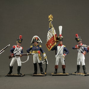 Puede incluir: Figuras en miniatura de soldados con uniformes históricos. Las figuras están pintadas con detalle, con detalles en azul, blanco, rojo y dorado. Una figura sostiene una bandera con un escudo. Los soldados están sobre pequeñas bases rectangulares.