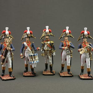 May include: Five miniature figurines of soldiers in historical uniforms. Each figure wears a bicorne hat with red and white accents, a blue jacket, and white pants. They hold various instruments, including a drum, trumpet, and clarinet.