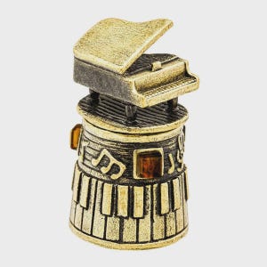 Może przedstawiać: Metalowy naparstek w kolorze złotym i czarnym z miniaturowym fortepianem na górze. Cylindryczny korpus ozdobiony jest wypukłymi nutami oraz wzorem klawiatury u podstawy, z akcentami w postaci małych, kwadratowych kamieni w kolorze bursztynowym.