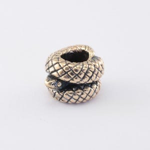 May include: A metallic bead shaped like a coiled snake with a textured, cross-hatched scale pattern. The bead features a bronze-toned finish with darkened recessed areas between the scales, highlighting the detailed serpent head and body.
