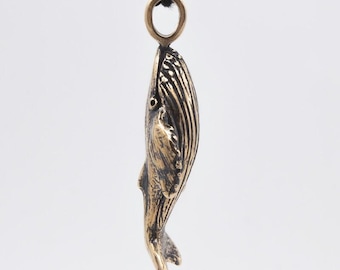 May include: A bronze-colored whale pendant suspended from a black cord. The pendant is detailed with textured lines and a small eye, showcasing the whale's form. The pendant is a nautical-themed accessory.