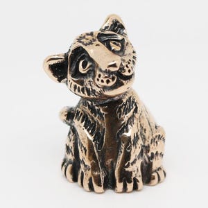 Op de afbeelding: Een bronzen beeldje van een zittende kat met gedetailleerde vachttextuur. De kat heeft grote ogen en een licht gedraaide kop, waardoor hij een nieuwsgierige uitdrukking heeft. De sculptuur is gemaakt van een metaalachtig materiaal met een donkere, verouderde afwerking.