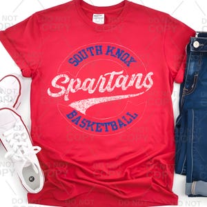 May include: A red t-shirt featuring the text "SOUTH KNOX Spartans BASKETBALL" in a circular design. The text is in white and blue, with a distressed, vintage look. The shirt is paired with blue jeans and white sneakers.