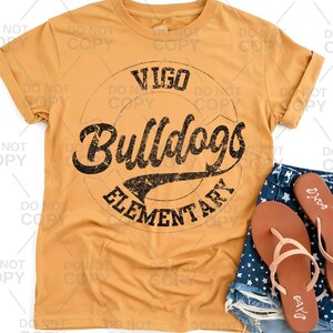 May include: Mustard yellow t-shirt with rolled sleeves. The shirt features a distressed black graphic with the words "Vigo Bulldogs Elementary" in a circular design. A pair of brown sandals and denim shorts are visible.
