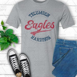 May include: A gray t-shirt with the words "Tecumseh Eagles Harrison" in a circle design. The word "Eagles" is in red script, and the other words are in blue. The shirt is paired with blue jeans and black and white sneakers.