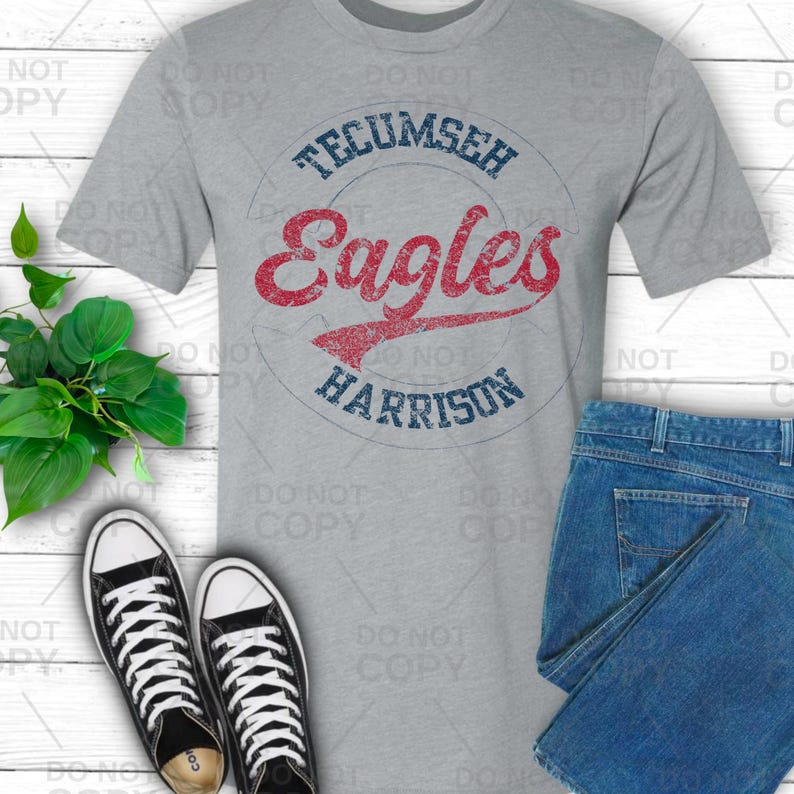 May include: A gray t-shirt with the words "Tecumseh Eagles Harrison" in a circle design. The word "Eagles" is in red script, and the other words are in blue. The shirt is paired with blue jeans and black and white sneakers.