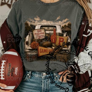 May include: A dark gray t-shirt featuring a graphic of a vintage truck bed filled with football gear, including a football, a foam finger with the number 1, a pennant reading GAME DAY, and a sign that says GO Princeton Tigers.