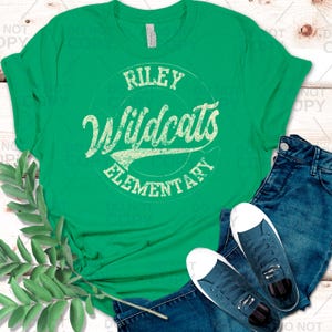 May include: A green t-shirt with the text "Riley Wildcats Elementary" in a distressed, vintage style. The shirt is paired with blue jeans and navy blue sneakers with white soles, creating a casual, everyday look.