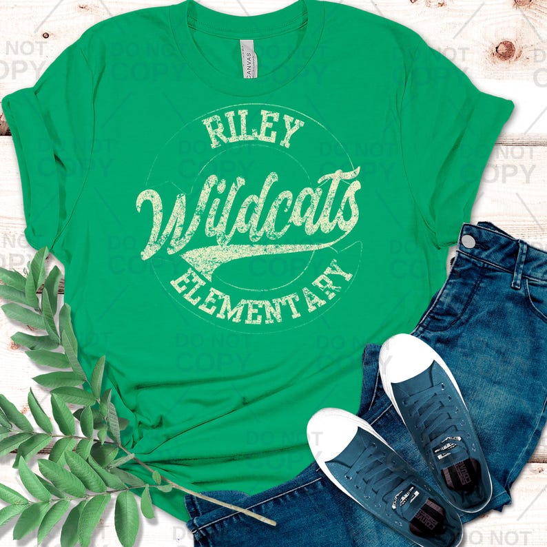 May include: A green t-shirt with the text "Riley Wildcats Elementary" in a distressed, vintage style. The shirt is paired with blue jeans and navy blue sneakers with white soles, creating a casual, everyday look.
