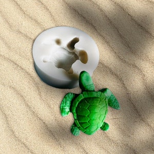 May include: A green turtle figurine next to a white silicone mold in the shape of a turtle. The turtle has a textured shell and is on a sandy surface. The mold is round.
