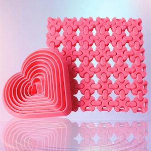 May include: A set of pink heart-shaped cookie cutters and a matching decorative mat. The cookie cutters are nested, with each one slightly smaller than the last. The mat has a repeating geometric pattern.