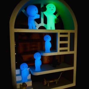 May include: A white, multi-tiered shelf unit with an arched top, displaying several glowing figurines. The figurines are in blue and green, with simple, rounded forms. A yellow duck and a small cat figurine are also present.