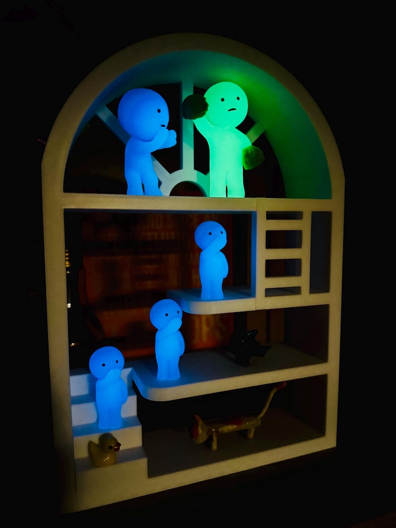 May include: A white, multi-tiered shelf unit with an arched top, displaying several glowing figurines. The figurines are in blue and green, with simple, rounded forms. A yellow duck and a small cat figurine are also present.