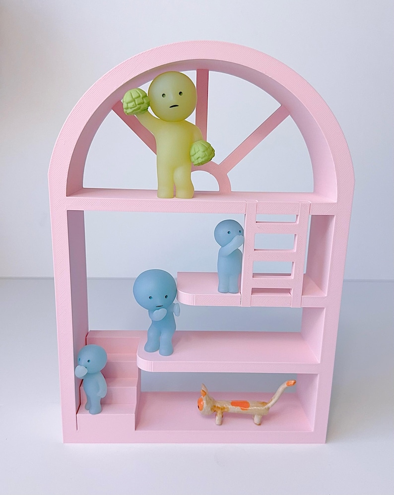May include: A pink, multi-tiered structure resembling a doll's house, with a curved top and several small, stylized figures. A yellow figure holds green objects. Blue figures are on the levels. A small cat figurine is also present.