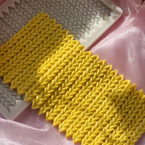 May include: Yellow, braided-patterned silicone mold with several yellow, braided-patterned bars. The mold is white. The background is pink and has a shiny, glittery appearance. The image is well-lit and the colors are vibrant.