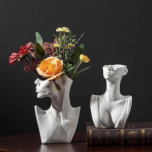May include: Two white ceramic vases shaped like busts with a floral arrangement in one vase. The flowers are yellow, orange, red, and green. The vases are on a wooden surface with a book underneath one of the vases.