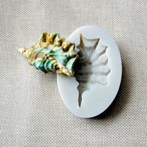 May include: A colorful seashell next to a white silicone mold. The seashell has shades of green, brown, and tan. The mold is oval-shaped with a detailed seashell impression. The background is a light beige fabric.