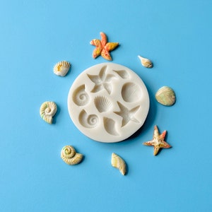 May include: A white silicone mold with seashell and starfish shapes, surrounded by several painted seashell and starfish decorations. The seashells and starfish are painted in shades of yellow, orange, and green. The background is a solid light blue.