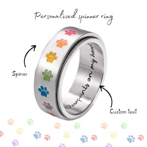 May include: A silver spinner ring with colorful paw prints and the text "Pawprints on my heart." The ring has the words "Personalized spinner ring" written above it. The ring is a pet-themed accessory.