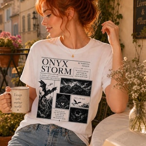 May include: White t-shirt with the text "ONYX STORM" and a graphic of a dragon and mountain scene. The shirt also features the text "A STORY OF COURAGE, A WAR FOR THE FUTURE" and "FLY...OR DIE." A beige mug with the text "Escape into another world" is held in the hand.
