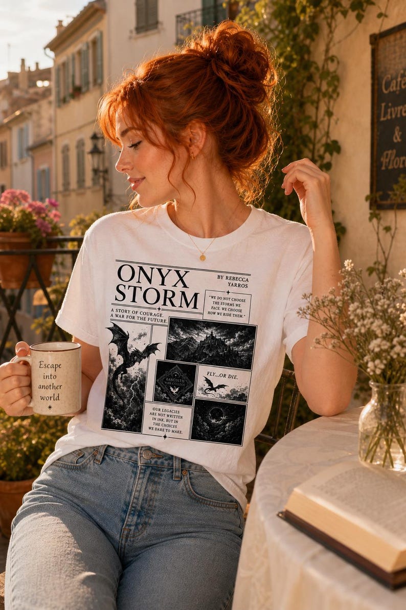 May include: White t-shirt with the text "ONYX STORM" and a graphic of a dragon and mountain scene. The shirt also features the text "A STORY OF COURAGE, A WAR FOR THE FUTURE" and "FLY...OR DIE." A beige mug with the text "Escape into another world" is held in the hand.