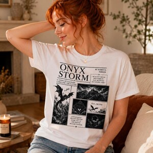 May include: White t-shirt with the text "ONYX STORM" and illustrations of a dragon, castle, and other fantasy elements. The shirt also includes the text "A STORY OF COURAGE, A WAR FOR THE FUTURE" and "FLY...OR DIE."