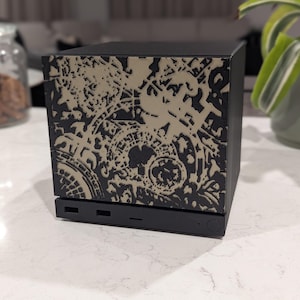 May include: A black square electronic device featuring a front panel with a beige, intricate, abstract gear-like pattern. The base of the unit includes two rectangular ports, a thin horizontal slot, and a circular power button on the right side.