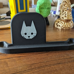 May include: A black plastic phone stand with a white cat logo. The stand has a small lip on the front and a larger lip on the back.
