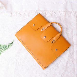 May include: A tan leather briefcase with a top handle and multiple pockets. The briefcase is on a white textured surface.