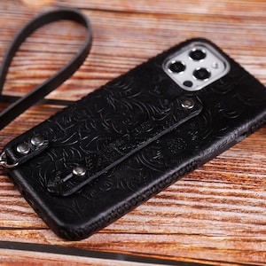 May include: Black leather phone case with a floral embossed design. The case has a wrist strap attached with silver-tone hardware. The phone case is designed to fit a smartphone.