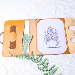 Op de afbeelding: Een bruin leren portfolio met een bruine leren flap en een tekening van bloemen in een vaas erin. De portfolio heeft een zak aan de linkerkant en een flap aan de rechterkant met drie metalen drukknopen.