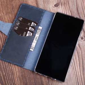 Puede incluir: Una funda de teléfono de cuero azul con una solapa que se abre para revelar ranuras para tarjetas de crédito y un bolsillo para efectivo. La funda está diseñada para sostener un teléfono inteligente.