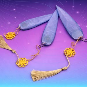 Puede incluir: Tres colgantes alargados de color azul claro con llaveros, cada uno adornado con un dije amarillo en forma de flor y una borla beige. Los colgantes están dispuestos sobre un fondo degradado púrpura y azul.