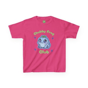 May include: A vibrant pink t-shirt with a cartoon frog graphic. The frog is light blue with big eyes and a small leaf. The text "Chubby Frog Club" is above and below the frog in green. Four blue stars surround the frog.