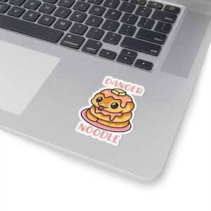 May include: A sticker featuring a stack of pancakes with a cute face, syrup, and a pat of butter. The sticker has the words "DANGER NOODLE" in pink text. The sticker is on a laptop.