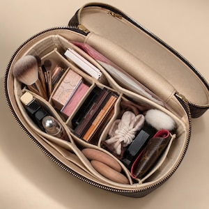 May include: An oval-shaped brown monogram cosmetic case with a gold zipper and a beige interior. The case features multiple internal dividers organizing various makeup items, including brushes, compacts, lipsticks, and a small pouch.