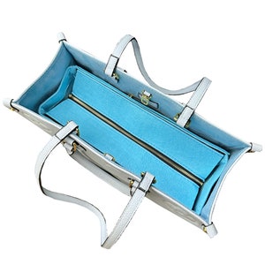 Peut inclure: Un sac cabas en cuir blanc avec une doublure zippée bleu clair. Le sac a deux poignées supérieures et une fermeture à glissière.