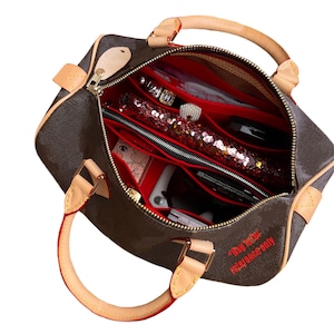 May include: A brown leather handbag with a red interior and multiple compartments. The bag is open and the interior is visible. The bag has a zipper closure and a top handle. The bag is labeled "*Bag Insert* Reference Only".