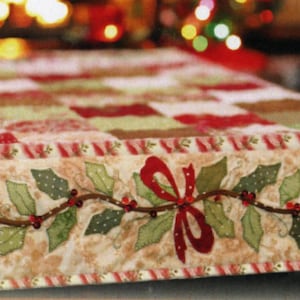 May include: A festive Christmas table runner with a patchwork design in red, green, and cream. The border features a holly and berry garland with a red bow. The runner is approximately 60 inches long.
