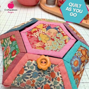 May include: A Blossie Basket, a small, hexagonal fabric container with a floral button closure. The basket features a variety of floral patterns in pink, blue, and orange. The text "QUILT AS YOU GO" is in a teal hexagon. The text "BLOSSIE BASKET" is at the top.