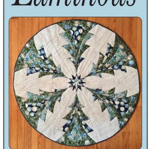 May include: A circular quilt with a starburst design in shades of blue, green, and white. The quilt is made with a variety of fabrics and has a quilted texture. The quilt is titled "Luminous" and is by Quilted Garden Designs by Jenice Belling.
